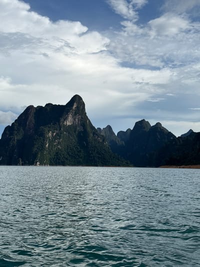 This landscape photograph depicts a scenic view of Khao Phang, Thailand. The image is oriented vertically, with the sky occupying the left side and a body of water the right. Towering, dark green, tree-covered mountains dominate the center of the frame, rising dramatically from the water's edge. Their rugged, rocky faces are visible in places, contrasting with the dense vegetation. The sky above is a bright blue, filled with fluffy white clouds that cast shadows on the mountains. The water in the foreground is a deep blue-gray, with a textured surface that suggests movement, possibly ripples or small waves. The scene evokes a sense of natural beauty and tranquility. The time of day appears to be daytime, with bright sunlight illuminating the scene. The weather seems clear with scattered clouds, typical of a tropical climate. There is no visible text in the image.