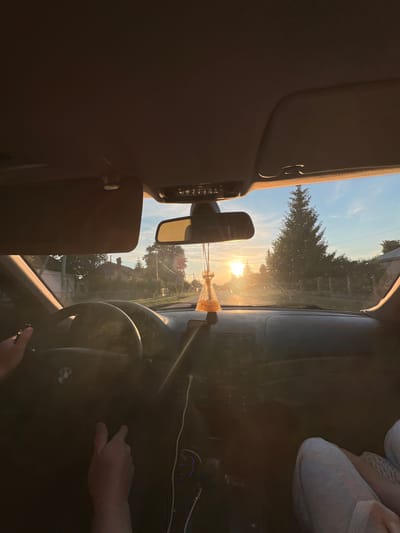 The image captures the interior of a car, specifically a BMW, at sunset in Velyki Hai, Ukraine. The perspective is from the passenger seat, looking forward through the windshield. The setting sun is directly ahead, creating a bright orange glare on the horizon and illuminating dust particles on the windshield.

In the foreground, the dark dashboard and a steering wheel with a BMW logo are visible. The driver's hands are on the steering wheel; the left hand is positioned at the top, and the right hand is lower. A sun visor on the driver's side is lowered, while the passenger-side visor is retracted. A rearview mirror hangs centrally, from which a yellow, pouch-shaped car air freshener with a tassel is suspended. To the right, below the windshield, a car charger with blue illuminated ports is visible, with white cables extending from it. The lower right portion of the image shows the legs of a passenger, dressed in light-colored pants.

Through the windshield, a rural or suburban road stretches into the distance. Houses with visible roofs and fences line both sides of the road. Numerous trees, including a prominent, tall evergreen tree on the right side, are present. Utility poles and overhead lines are also visible along the road. The sky above is a clear blue, transitioning to warm orange and yellow hues near the horizon due to the sunset.