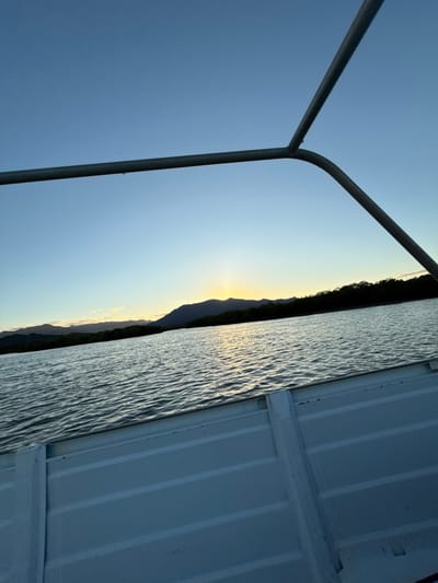 The image presents an outdoor waterscape at either sunrise or sunset, observed from a boat. The lower foreground depicts the interior of a boat, characterized by light-colored, horizontally ribbed panels comprising the hull or seating structure. A metallic, tubular frame, likely a canopy support, extends horizontally across the upper portion of the image, then curves and descends diagonally on the right. A single water droplet is suspended from the underside of the horizontal tube, approximately centered.
The midground features a body of water exhibiting moderate surface ripples. This water body reflects a distinct, vertical column of bright yellow-orange light emanating from the horizon. The water's surface appears dark blue-grey, marked by areas of illuminated reflection.
The background is composed of a dark, linear band of dense green foliage, consistent with trees or mangroves, bordering a distant landmass. Behind this vegetation, a continuous range of dark, undulating mountains or hills stretches across the entire horizon. The sun is positioned low on the horizon, slightly to the left of the image's vertical center, emitting a concentrated yellow-orange glow upwards. The sky above is clear, transitioning from a deep blue at the top of the frame to lighter blue and yellow-orange hues near the horizon line.
This scene is located within the city of Dumbéa, France.