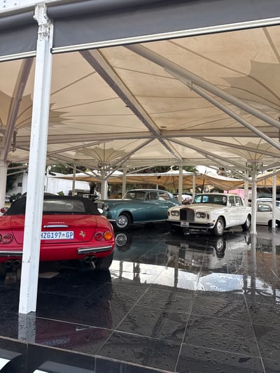 The scene depicts several vintage cars parked under a sheltered area, likely in Kraaifontein, South Africa. The foremost car is a red convertible, with a license plate reading "HZG197 GP." Behind it are vehicles in shades of teal and white. The cars are parked on a black tiled surface that appears wet. The sheltered area features a white framework supporting a light-colored canopy overhead. The image is taken from a low angle, looking upwards towards the vehicles and canopy.
