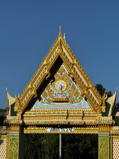 This is a detailed, full-frame, eye-level, medium shot of the ornate, gilded entrance of a temple in Baan Tha Po, Thailand. The temple's architectural style is unmistakably Thai, characterized by its steep, triangular roofline adorned with intricate gold leaf detailing and decorative motifs. The roof is crowned with pointed finials, and its edges are lined with a repeating pattern of gold ornamentation.

Dominating the upper portion of the entrance is a central, circular emblem featuring a silver Buddha statue seated in meditation, encircled by elaborate gold scrollwork against a vibrant blue background. Below this, a horizontal band of gold relief work runs across the structure, separating the upper decorative elements from the lower portion of the entrance. The lower section displays more detailed gold embellishments and what appear to be Thai characters rendered in blue script.

The entrance itself is framed by columns and archways, also decorated with gold and green patterns. The green elements often feature a swirling, foliate design, while sections are filled with a white diamond-shaped mesh pattern. The background reveals lush green foliage, suggesting the temple is situated amidst a natural environment, and a clear, deep blue sky. The lighting indicates it is daytime, with strong sunlight casting shadows, highlighting the three-dimensional quality of the intricate carvings. No people are visible in the frame.