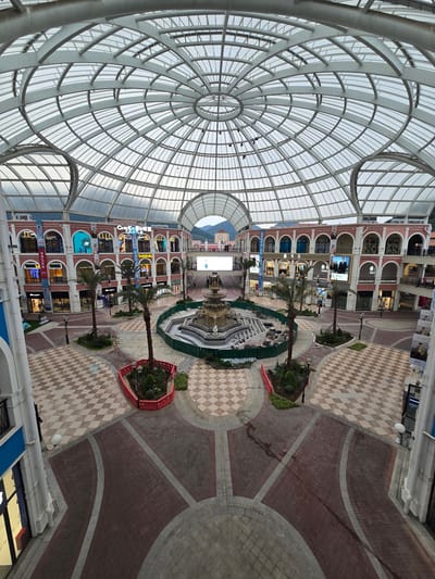 The image shows an interior view of a shopping mall with a large, domed glass roof. The mall features multiple levels of shops with arched entrances and balconies. In the center of the mall is a fountain surrounded by planters and palm trees. The ground is paved with a patterned tile design. In the background, mountains are visible through the open sections of the roof. The scene is brightly lit by natural light filtering through the glass and artificial lighting from the shops. There are no people visible in the image. The overall impression is that of a modern, spacious, and well-maintained commercial center. This location is likely the "Fuyang International Castle" in Fuyang, China, a shopping mall known for its distinctive architecture.
