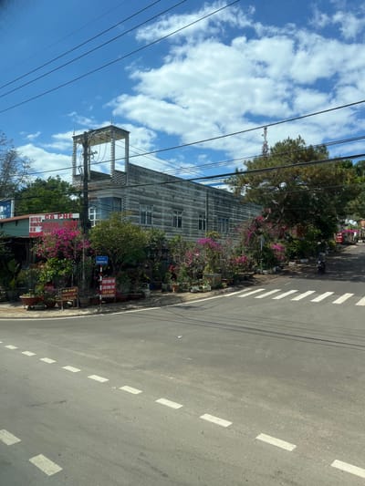 A street scene shows a tall, weathered building with an open tower-like structure. Bougainvillea vines add color along the sidewalk. A utility pole with multiple wires stands next to the building. The road has painted white dashes. The sky is blue with scattered clouds. Green trees are also visible.