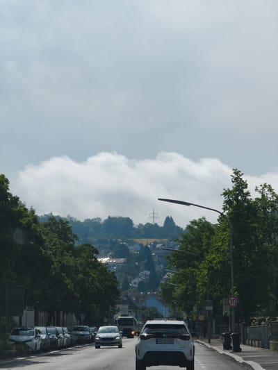 The image depicts a street scene in Lörrach, Germany. The perspective is from a vehicle traveling down a two-lane road. On the left side of the road, there is a row of parked cars and a sidewalk lined with trees. On the right side of the road, there are more trees, streetlights, trash cans, and a speed limit sign indicating 30 km/h. In the distance, the road leads into a residential area with houses nestled on a hillside. The sky is partially cloudy with a band of thick, white clouds obscuring the upper portion of the view. A power pylon is visible in the background. The overall lighting suggests it is daytime, possibly a slightly overcast day.