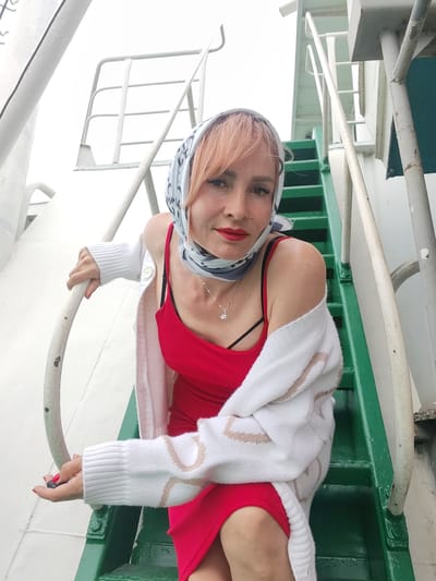 A woman with light pink hair, wearing a red dress and a white cardigan, poses on a set of green metal stairs with white handrails. She has a patterned scarf tied around her head, covering her hair. Her makeup includes red lipstick and eyeliner. She is also wearing a silver necklace with a small pendant. Her left hand is resting on a white handrail, and her right hand is holding a dark object, possibly a phone. The stairs appear to be on a boat or ship, indicated by the white metal structures and the presence of railings. The background shows more of the white structure and the sky, suggesting an outdoor setting. The lighting indicates it is daytime, and the overcast sky suggests cloudy weather. The woman's expression is a slight smile, and she appears relaxed and confident. No visible text is present in the image. The location context is Bereg, Russia.