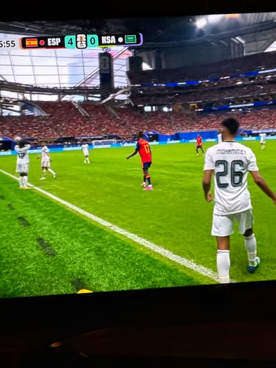 The image captures a moment during a soccer match, likely being broadcast on a television. A scoreboard in the upper left corner displays "5:55" and indicates a score of "ESP 4" against "KSA 0". Several soccer players are visible on a green field. One player in the foreground is wearing a white jersey with the number "26" and the name "MOHAMMED" on the back. Another player in a red jersey with the number "17" is in the mid-ground. The stands of a stadium, filled with spectators in red, are visible in the background. The lighting suggests an indoor or evening setting with artificial illumination. The image is a photograph of a screen, with a dark border at the bottom. There is no visible connection to the city of Bykovo, Russia.