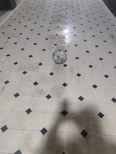 The image displays a single, heavily used soccer ball resting on a textured, tiled outdoor surface in Rishtan, Uzbekistan. The ball is dirty, scuffed, and appears partially deflated, featuring remnants of white, green, and silver-grey panels. The ground is composed of light-colored, square tiles with a granular or stippled surface pattern. Dark, diamond-shaped tiles are systematically inlaid at the intersections of the larger tiles, forming a consistent grid. The tiling exhibits general dirt and wear. A distinct, bright reflection is present on the tiles in the upper mid-ground, indicating a localized light source. A large, dark, indistinct shadow, consistent with a human silhouette, extends across the lower right portion of the scene. A dark, vertical architectural element with a textured surface is visible along the left edge, adjacent to a lighter, raised curb or step. A rectangular utility access panel or drain cover is embedded within the tiled surface further into the background. No individuals are directly visible.