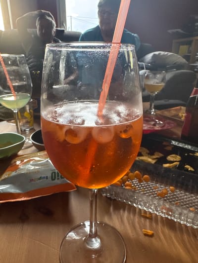 A high-angle, indoor shot shows an Aperol spritz in a wine glass with a pink straw, next to a clear plastic tray with small orange snacks and a bag of Lupilu sticks on a wooden table. In the background, blurred figures of people are visible, along with other glasses and objects. The location is Baillonville, Belgium.