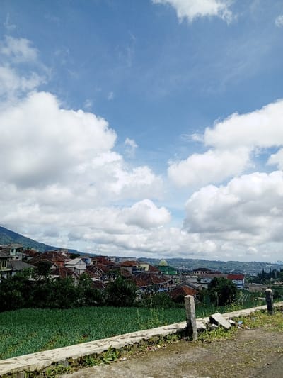 A daytime scene reveals a panorama of structures beneath a sky filled with fluffy white clouds. Green foliage and fields fill the immediate foreground. A concrete barrier runs along the edge of a road, separating it from the vegetation. Beyond the fields, houses with red-tiled roofs and other buildings cluster together on rolling hills, rising towards a distant mountain range visible on the left.