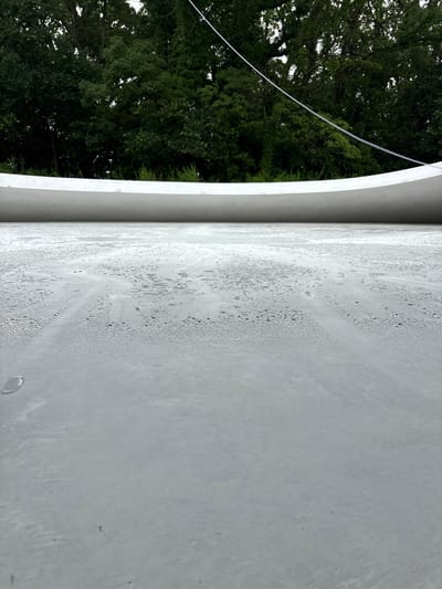 The image is a low-angle shot of a wet, gray surface, possibly concrete or asphalt, with numerous small water droplets and a few larger puddles. In the background, a curved, white structure runs horizontally across the frame, partially obscuring a dense line of dark green trees. A thin, white wire or cable is visible diagonally against the trees. The overall impression is one of an outdoor, possibly urban or park-like setting, after or during rainfall. The scene is devoid of any people or specific landmarks that would indicate its location within Tonosho, Japan.