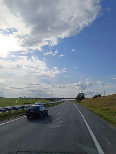 This is a frontal view of a highway in Пухавіцкі сельскі Савет, Belarus, taken from inside a moving vehicle. The sky is a mix of bright blue with scattered clouds, some of which are dark and heavy, suggesting an impending weather change. Sunlight filters through gaps in the clouds, casting rays onto the scene. The highway is paved with asphalt and has clear white lane markings. On the left side of the road, a metal guardrail separates the highway from a flat green field with some sparse vegetation and what appears to be industrial structures or smoke in the distance. On the right side, a sloping grassy embankment rises, with some bushes and trees. A dark-colored Audi station wagon is driving in the same direction as the camera vehicle, positioned in the left lane. Another white van is visible further ahead in the right lane. An overpass or bridge crosses the highway in the distance. Electrical power lines and poles are visible on the right embankment. The overall impression is a typical highway scene on a partly cloudy day.