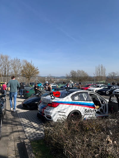 The image depicts a crowded outdoor parking area on a clear, sunny day in Meuspath, Germany. Numerous high-performance and race-modified automobiles are parked, alongside approximately 20-30 individuals observing the vehicles and interacting.
In the foreground, a heavily modified white BMW M3-series sedan (F80) is prominent on the right, partially obscured by dark, leafless bushes. This vehicle features extensive track modifications, including a large black rear wing with red accents, a visible roll cage through its open driver's side door, and a white livery covered with "BIMMER TUNING," "NANKANG," "RECARO" decals, and red, white, and blue BMW M-color stripes along its side. "Safety Official Car of Bil..." is visible on its side. A person in tan clothing is seated inside the vehicle. To the left of the BMW, a black, low-slung supercar, possibly a Lamborghini Huracan, is parked.
In the midground, a diverse collection of performance cars, including other BMWs (e.g., a dark green M4), Mercedes-AMGs, Porsches, and various sedans and coupes in colors such as red, white, black, and teal, are parked in rows across a paved surface composed of interlocking concrete pavers and asphalt.
Multiple individuals are distributed throughout the scene. On the far left foreground, a male with short brown hair, wearing a teal long-sleeved shirt and blue jeans, walks away from the camera. In the midground, groups of people are standing near the parked cars, conversing or observing. One man wearing a black t-shirt and cap is visible near the black supercar, facing towards the viewer. A woman with blonde hair, wearing a brown top, is seen interacting with a man near a red vehicle.
The background features a wire mesh fence separating the parking lot from a landscape of mostly bare deciduous trees and distant green hills under a bright, clear blue sky with minimal high-altitude clouds. Strong direct sunlight casts distinct shadows. Several utility poles are visible in the far background. The overall environment suggests an automotive enthusiast gathering or car meet, likely associated with the Nürburgring complex due to the vehicle types and location.