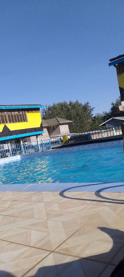 A swimming pool is in the foreground, with clear blue water and a tiled edge. Beyond the pool, a yellow building with blue trim is visible. In the background, there are trees and a glimpse of another building. The sky is clear and blue. The foreground features light-colored tiled flooring with shadows cast across it.