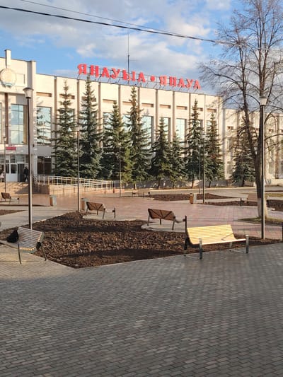 An outdoor, eye-level shot captures a plaza and a prominent building in Yanaul, Russia, likely on a clear day with some clouds. The large, light-colored building in the background features the city name "ЯНАУЛ • ЯНАУЛА" in red, block letters across its facade. Tall, narrow evergreen trees (possibly spruce or fir) are planted in front of the building, creating a green line between the structure and the plaza. The building's architecture appears functional, with large windows reflecting the sky.
The foreground and midground feature a paved plaza made of interlocking gray and reddish-brown bricks. Several modern-looking wooden benches with metal frames are scattered across the plaza. Garden beds, filled with dark earth or mulch, separate sections of the paving, suggesting recent landscaping or preparation for planting. One black bag is visible on a bench in the lower-left. Streetlights with dark poles are strategically placed throughout the area. To the right, a large deciduous tree with bare branches stands tall, indicating a season like late autumn, winter, or early spring. Overhead, utility wires cross the sky.
A ramp with white railings leads up to the entrance of the building. A few individuals can be seen near the entrance, either entering or exiting. The overall impression is one of a well-maintained public space, quiet but ready for activity. The light suggests it is daytime, with shadows indicating either morning or late afternoon sun.