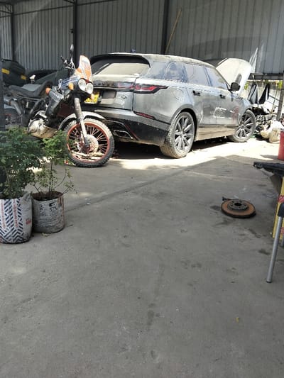 The scene is an interior or covered exterior space, likely a vehicle repair workshop, located in Mkomani ward, Kenya. The ground is a light grey concrete slab, showing wear and dust. The background features a corrugated metal wall.
In the left foreground, two plants are contained within cylindrical planters made from repurposed woven sacks, one displaying partial "CROWN" branding. Immediately behind these, an off-road style motorcycle is parked, facing slightly left. It features a dark frame, dual headlights, an orange windscreen/fairing with a "30" decal, and a front wheel with an orange-red rim and knobby tire.
Behind the motorcycle, a dark grey Range Rover Velar SUV is parked, facing slightly left. The SUV is visibly covered in a layer of dust. Its hood is open, indicating it is undergoing maintenance or repair, and the left rear passenger door is partially ajar. The rear of the vehicle displays "VELAR" and "ROVER" badging, along with a yellow Kenyan license plate. Multi-spoke dark alloy wheels are visible on the SUV.
To the far right, partially obscured, is a yellow and silver metallic support structure or piece of equipment. Further into the background on the right, a disorganized pile of various objects, possibly vehicle components or scrap, rests against the corrugated wall. On the concrete floor in the mid-right foreground, a single, visibly rusted disc brake rotor lies flat. No individuals are present in the depicted scene.