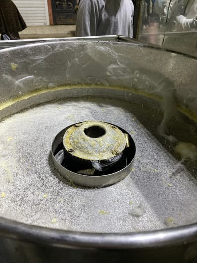 The image displays a close-up, slightly downward-angled view of a well-used cotton candy machine situated in Sidi Chahmi, Algeria. The machine's central spinning head is heavily encrusted with a thick layer of solidified yellow and beige sugar residue. The surrounding metallic bowl, likely stainless steel, contains a significant accumulation of fine white granular sugar, larger yellow-beige sugar clumps, and fibrous white cotton candy material clinging to its interior surface and base. In the blurred background, two individuals are partially visible: one on the left wears a dark, patterned garment, and another on the right wears a plain gray long-sleeved shirt. Behind them, a building facade with a closed white roller shutter and a dark, ornate wooden door is discernible, indicating an outdoor or semi-outdoor commercial environment under low light conditions.