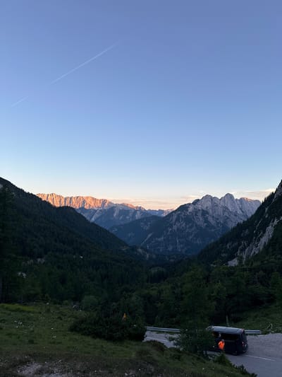 The image depicts a serene mountain landscape at dusk, with the sun casting a warm, golden glow on the jagged peaks in the distance. The sky is a clear, soft blue, with a faint airplane contrail stretching across it. The foreground is dominated by a lush, green forest that slopes down into a valley. A paved road winds through the scene, with a dark-colored van parked on the shoulder. A person wearing an orange shirt is visible near the open rear door of the van. The location appears to be in the vicinity of Soča, Slovenia, a region known for its stunning alpine scenery. The overall atmosphere is one of tranquility and natural beauty.