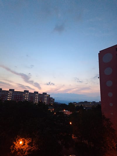 The image depicts a cityscape at dusk, likely in the city of Most, Czechia. In the foreground, dense, dark foliage of trees obscures the ground, with several illuminated streetlights casting an orange glow. Beyond the trees, a long, multi-story apartment building is visible, its windows lit, indicating occupancy. The building is characterized by its repetitive rectangular windows and a facade with red and white sections. Further in the distance, another row of apartment buildings appears, also illuminated. The sky above transitions from a pale blue near the horizon to a deeper twilight blue overhead. Wispy clouds are present, tinged with soft pink and orange hues from the setting sun. To the right of the frame, a tall, red-colored building with large, circular, light-colored decorative elements is prominent, its top portion cut off by the frame. The overall impression is one of an urban residential area as day transitions into night.