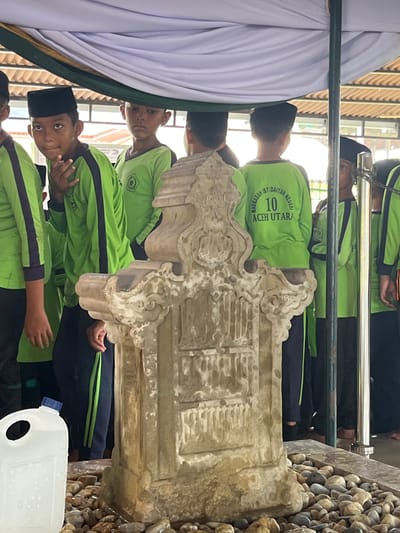 A group of young boys, all wearing black peci caps and bright green long-sleeved shirts with dark blue accents, are gathered around an ornate stone monument. The monument, located at the foot of what appears to be a grave, is heavily weathered and decorated with intricate carvings. The boys, some looking towards the camera and others away, stand partially obscured by a draped white and green fabric canopy. Behind them, the environment suggests an outdoor setting with glimpses of a building's roof and what could be a metal fence. In the foreground, to the left, a clear plastic water jug with a blue lid is positioned among a scattering of smooth, grey and brown stones. The scene appears to be at a place of historical or religious significance, possibly a cemetery, in the city of Beuringen, Indonesia. The attire of the boys suggests they might be students on an outing or participating in a commemorative event.