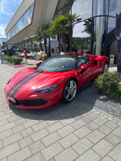A red Ferrari 296 GTS convertible is parked on a paved walkway in front of a building. The car has black racing stripes and silver wheels with yellow brake calipers. The license plate reads "ZH-UB 63". The building features glass windows reflecting palm trees and the sky. A banner with the words "SUMMER VIBES" is visible on the building. A small bush sits next to the rear of the car. In the background, there is a table with balloons, a small group of people, and some retail displays. The scene appears to be in Jestetten, Germany, based on the license plate.