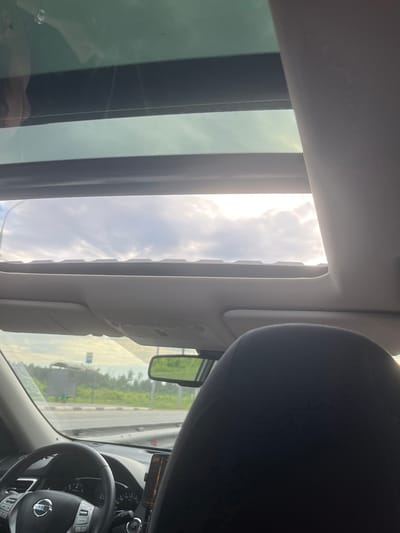The image shows an interior view from within a car, specifically oriented towards the partially open sunroof. The sky is visible through the sunroof, with a mix of clouds and sunlight. A rearview mirror and parts of the dashboard and steering wheel are also visible. Outside the car window, greenery and a road can be seen. While the image does show elements that could be associated with a road or city, there is insufficient information to confidently assert that the location is Monino, Russia. There are no identifiable people or specific actions occurring within the frame.