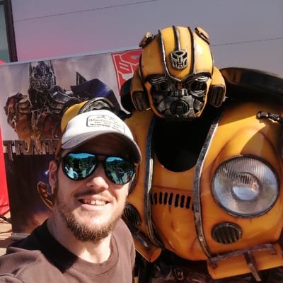 This image, taken in Трубачеевка, Russia, depicts a male individual taking a close-up selfie with a large, weathered yellow statue resembling Bumblebee from the Transformers franchise. The man, positioned on the left side of the frame, has a light complexion, a trimmed beard, and is smiling broadly towards the camera. He wears a dark brown long-sleeved shirt, a white baseball cap with a 'Montana 1972 Outdoor' patch, and blue mirrored sunglasses that reflect an outdoor scene with multiple people and green foliage.
The Bumblebee statue, occupying the right and center of the image, is predominantly yellow with metallic, distressed accents, and features an Autobot symbol prominently displayed on its forehead. Its design incorporates elements of a classic Volkswagen Beetle, evident in the round headlight on its lower right side and the grille-like vents.
In the background, a large poster for the Transformers franchise is visible. It features a depiction of Optimus Prime and the partially visible 'TRANSFORMERS' logo, along with another red Autobot symbol. The overall setting appears to be an outdoor public event or display area, under bright daylight.