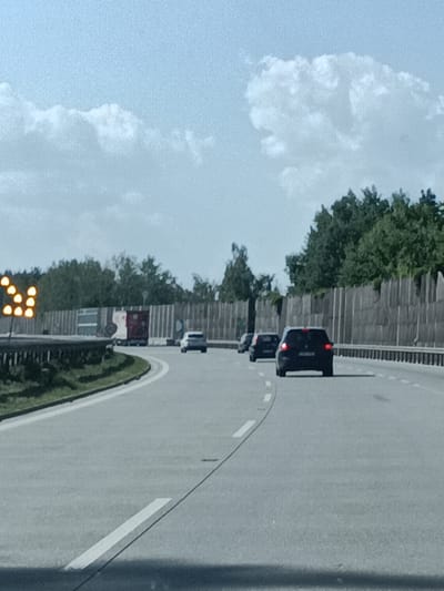 The image shows a highway with several cars. A dark-colored SUV is in the foreground, followed by another dark-colored car. In the distance, there is a white car. On the left side of the highway, there is a series of illuminated orange arrows pointing to the left. Beyond the highway, there are trees and a tall, grey sound barrier. The sky is partly cloudy. The image appears to have been taken from a moving vehicle, suggesting the highway is in use. The specific location within Lensedly, Czechia, is not discernible from the visual information provided.