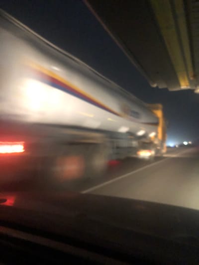 The image captures a motion-blurred view of a white train, likely a high-speed or commuter train, moving past the viewer in a dimly lit environment, possibly at night or indoors like a station or tunnel. The photograph is oriented vertically, with the train appearing to stretch along its length.
The train itself is predominantly white, with a prominent livery consisting of colorful stripes—yellow, red, and blue—running horizontally along its side. The motion blur is significant, especially on the train, indicating considerable speed. Bright red lights, possibly taillights or warning lights, are visible towards the rear section of the train, glowing intensely against the dark background.
The surrounding environment is dark and appears to be composed of structural elements. On what would be the left side of the train (if viewed upright), there are some metallic or yellowish structures, possibly overhead gantries or parts of a station platform, along with a dark, shadowy expanse. On the opposite side of the train, a dark, vertical wall or structure is visible, which could be a tunnel wall or another platform edge. Faint, distant lights can be seen in the background, adding to the nighttime or indoor impression.
The perspective suggests the photo was taken from a vantage point slightly above and to the side of the moving train, looking down its length as it passes. No discernible text or logos are clearly visible on the train due to the motion blur and lighting conditions. The overall mood is dynamic and fast-paced, conveyed by the streaking lights and blurred lines.