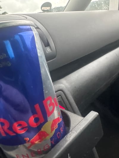 The image provides an angled, close-up view of a car's interior, likely in Schopp, Germany, during daylight on an overcast or rainy day, as indicated by water droplets on the window. The primary subject is a blue and silver Red Bull energy drink can, prominently placed in a storage slot or cup holder within the car's console area.
On the left, a light grey car door panel features a black, textured rotary dial marked with the number "0," likely a control for mirrors or lighting. Below this, a section of a speaker grille is visible. Through the adjacent car window, a blurred circular object, appearing to be a side mirror, reflects a hazy outdoor scene.
The car's central console and surrounding surfaces are dark grey or black plastic, showing some minor dust or wear. A faint red light can be observed emanating from deeper within the console, possibly an indicator. The Red Bull can clearly displays its red "Red Bull" logo with the charging bull graphic, and the words "ENERGY DRINK" are partially visible. There are no people in the image, and the scene depicts a common roadside or travel context where an energy drink is conveniently stored.