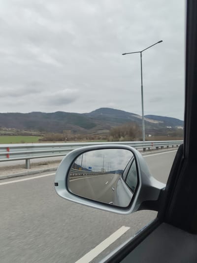 The image shows a view from inside a moving car, likely on a highway. The side mirror of the car prominently features the road behind, stretching into the distance with lane markings and light poles. The side mirror also reflects a view of the road ahead, with other cars visible in the distance. Outside the car window, there is a highway barrier and a landscape featuring a low mountain range under an overcast sky. The location is identified as Novachene, Bulgaria.