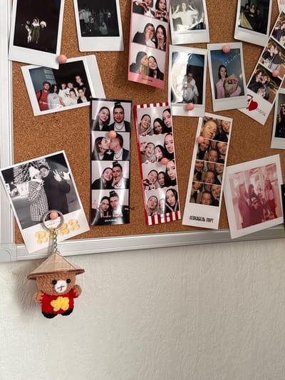 A corkboard, likely in an interior setting, is populated with photos and a small bear trinket. The photos appear to be a mix of instant prints and photobooth strips, depicting various people in what seems like candid or casual poses, possibly friends or family. Some photos are affixed with pushpins, adding to the collage-like aesthetic. A photobooth strip includes the text "СЕВКАБЕЛЬ ПОРТ" in Russian, indicating that the photos were likely taken at Sevkabel Port, a public space in Severnyi, Russia. A small, handmade bear with a conical hat, attached to a keyring, is pinned to the board, adding a personal touch. The background is a plain white wall, which contrasts with the cluttered and personal nature of the corkboard display. The overall scene suggests a collection of memories and personal mementos displayed in a casual manner.