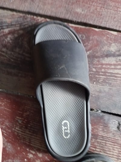 A single black slide sandal rests on a weathered wooden surface. The sandal has a textured, light gray footbed with a circular embossed logo featuring two interconnected C-shapes. The outer sole is also black. The wooden surface appears to be made of planks with visible grain and some wear. The lighting suggests it could be daytime. There is no discernible text on the sandal other than the logo. The overall scene is simple and focuses on the sandal against the rustic background.