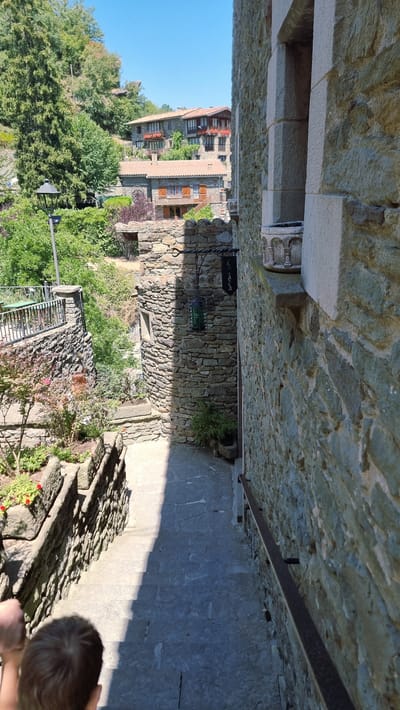 The image depicts a narrow cobblestone staircase winding down between stone buildings in Rupit i Pruit, Spain. The architecture is characteristic of a medieval village, with rough-hewn stone walls and terracotta-tiled roofs visible in the background. Lush green trees and foliage flank the staircase, adding a touch of nature to the scene. Sunlight casts a strong shadow across the stairs, indicating it's a bright, sunny day. A railing made of dark metal runs along the right side of the stairs for support. In the foreground, the back of a child's head is visible, suggesting they are walking down the stairs. A hanging lantern with a green glass shade adorns the stone wall on the left. The overall atmosphere is one of old-world charm and tranquility.