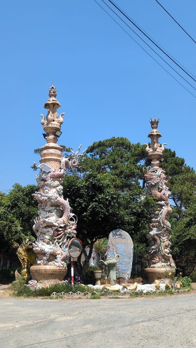 The image shows two ornate, dragon-covered pillars located in Lâm Sơn, Vietnam. The pillars are made of a light-colored material and are adorned with colorful ceramic dragons. They are positioned alongside a road and surrounded by lush greenery and trees. The sky is clear and blue, with power lines visible in the upper left corner. A weathered sign with blue characters is visible between the pillars. Various objects such as a mirror, a pot, and what appears to be a solar panel are incorporated into the display.