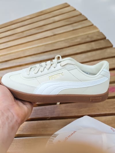 A person's hand is holding a light cream-colored Puma sneaker. The sneaker has a white stripe on the side and a brown rubber sole. The Puma logo is visible in gold on the side of the shoe and embossed on the heel of the sole. The background consists of a wooden slatted surface, possibly a table or bench, with a light-colored, out-of-focus object in the foreground. There is no discernible information within the image to suggest a location in Nola, Italy.