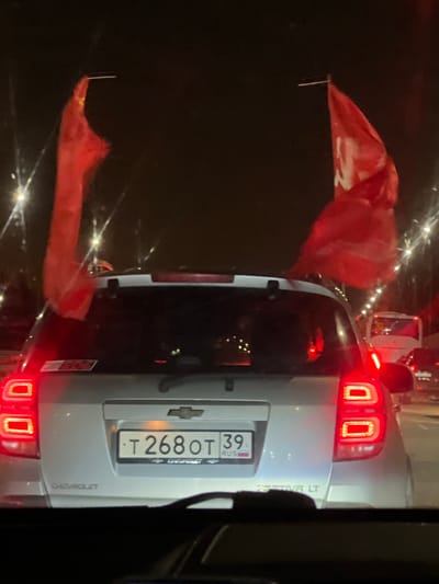 A night-time close-up shot of the rear of a silver Chevrolet Captiva LT with a Russian license plate "T268OT 39 RUS". Two red flags are affixed to the car, one to the left of the left tail light and one to the left of the left side of the rear windshield. The location is Зеленая Роща-1 Киз, Russia. The scene appears to be taken from another car, with a portion of the car visible on the right side of the frame. The background is dark with lights visible.