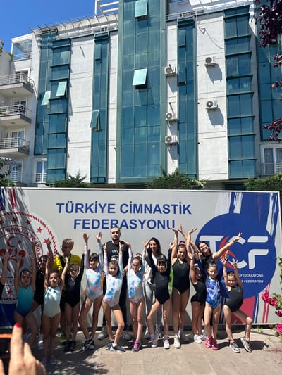 In Sancaktepe, Turkey, in front of a modern building with multiple stories and blue-tinted windows, a group of young gymnasts stands with their hands raised in a celebratory pose. The building's facade features several air conditioning units and balconies. A large banner behind the gymnasts displays the text "TÜRKİYE CİMNASTİK FEDERASYONU," which translates to "Turkish Gymnastics Federation." The banner also includes the logo of the federation, a stylized "TCF" within a blue circular emblem. The gymnasts are wearing various leotards, some plain and others with colorful patterns. Two adults, likely coaches, are positioned behind the children, also participating in the pose. The ground is paved with light-colored tiles, and to the right of the image, a pink flowering bush is visible. The sky is clear and blue, indicating a sunny day.