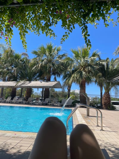 The image captures an outdoor swimming pool scene from a first-person perspective, looking across the pool. In the foreground, the partially visible knees and thighs of a person suggest they are relaxing by the pool, possibly with their feet in or near the water.

The upper portion of the image is dominated by a clear, bright blue sky, indicating sunny weather. Lush green foliage, featuring small pink flowers, frames the left side of the view. Several prominent palm trees with substantial trunks and green fronds are scattered around the pool area, their leaves swaying gently.

The swimming pool itself is filled with vibrant blue water. A metallic handrail leads into the pool on the right, and a stream of water is actively flowing into the pool, creating ripples and splashes. The pool is surrounded by a tiled deck area. Along the far side of the pool, several lounge chairs are arranged under a shaded pergola or sunshade structure, suggesting a resort setting. In the background, beyond the pool and trees, a fence and some buildings are visible.

The time of day appears to be midday or early afternoon, characterized by bright sunlight and strong shadows. The overall scene evokes a sense of relaxation and vacation in a warm, sunny climate. There is no visible text in the image. The location context is Ayia Napa, Cyprus.