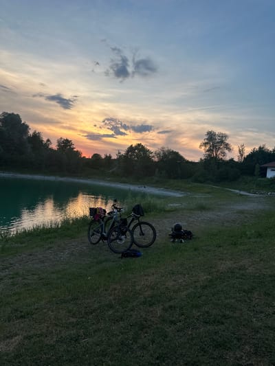 Two bicycles are parked on a grassy area next to a body of turquoise water. The sky is a gradient of blue and orange, with clouds scattered across it. The scene appears to be set at dusk or dawn. The bicycles are equipped with baskets and bags, suggesting they may have been used for a leisurely outing. In the background, trees line the shore, and a small building is visible in the distance. The overall impression is one of tranquility and nature. The image appears to have been taken in Halfing, Germany, given its picturesque landscape and the characteristic rural setting.