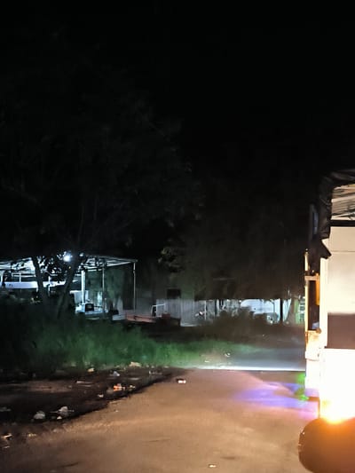 This night scene, set in Lhoknga, Indonesia, depicts a dimly lit outdoor area. In the foreground, a rough path or unpaved road is brightly illuminated by the headlight of a vehicle, a portion of which, appearing white or light-colored, is visible on the right side of the frame. The strong light casts distinct blue, purple, and green hues onto the ground directly in front of the vehicle. Scattered litter, including bottles and other debris, is visible on the ground.
A strip of green grass borders the path on the left. Beyond this, large, dark trees dominate the mid-ground and left side of the image, their branches silhouetted against the dark sky. Beneath the trees, several open-sided structures, possibly sheds or workshops with light-colored roofs and visible support beams, are faintly illuminated from within. A white fence or wall can be seen behind these structures on the right. The overall atmosphere suggests a utilitarian or industrial area after dark. No people or visible text are present.