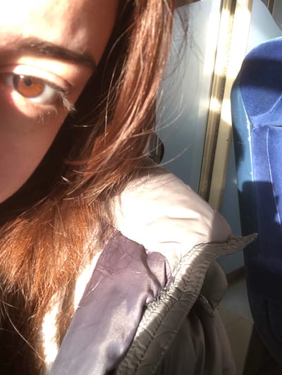 The image presents a close-up, angled view of an individual positioned inside a vehicle in Dettwiller, France. The left side of the subject's face is visible, featuring a brown left eye with a dark pupil and defined eyelashes, partially in shadow. Reddish-brown hair, illuminated by direct sunlight, frames the face and extends across the left side of the frame. The individual wears a light gray or lavender padded jacket, the upper portion of which shows a distinct textured seam. To the right, a section of a blue upholstered seat is visible, indicating an interior setting consistent with a train or bus cabin. Strong natural light enters from the right, casting bright highlights on the subject's hair and jacket and creating vertical light patterns on the internal window structure. The individual's gaze is directed forward, seemingly towards the camera.