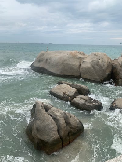 This image depicts a coastal scene at Cà Ná, Vietnam. The foreground is dominated by several large, weathered rocks partially submerged in choppy, greenish-blue water. Waves crash against the rocks, creating white foam. In the background, the vast ocean stretches to the horizon under a cloudy, overcast sky. A few distant ships and structures are visible on the water, suggesting maritime activity. The overall environment is natural and rugged, with the sea and rocks being the primary subjects. There are no visible people in the immediate foreground or mid-ground.