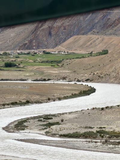 An aerial, wide-angle shot captures a wide, meandering river flowing through a dry, mountainous landscape. The river is a milky white color and takes up a significant portion of the foreground, with small islands and gravel bars visible within its flow. The banks of the river are lined with sparse, dry vegetation and rocky terrain.
In the middle ground, the landscape transitions to green agricultural fields, indicating a more fertile area. Several small, white-domed structures, likely stupas or chortens, are scattered amongst the fields. A small village with white-washed buildings and a prominent cell tower is nestled at the base of the mountains.
The background is dominated by steep, rocky mountains with visible layers of sediment, suggesting a geologically complex area. The sky is overcast and a dark green band is visible across the top of the frame, likely the edge of a banner or prayer flag. The image depicts a remote, high-altitude settlement in Zangla, India, characterized by its stark natural beauty and the presence of human habitation adapted to the harsh environment. There are no people or animals visible in the image.