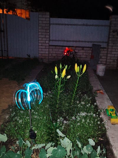 A night scene shows a garden area next to a brick fence and a grey metal fence. In the foreground, a blue fiber optic garden light illuminates the area. Behind it, a flower bed with green plants and small white flowers is visible. To the right, a yellow toy bulldozer sits on a concrete path. In the background, a building with illuminated windows is visible through a gap in the fence. The overall scene suggests a residential backyard in Kotovsk, Russia.