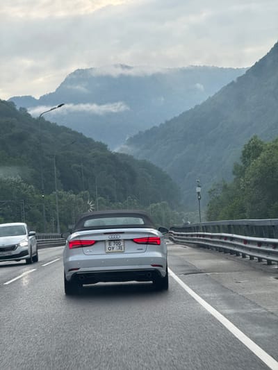 The image shows a white Audi convertible driving on a wet asphalt road with guardrails on the right side. The car's license plate is visible and reads "E 700 OY 35". In the background, there are lush green mountains with a cloudy sky. The setting appears to be a highway in a mountainous region, possibly the city of Chvizhepse, Russia, given the presence of a speed camera on a pole and the general landscape. The road is wet, indicating recent rain or mist. There is another white car in front of the Audi on the left lane. The overall atmosphere is overcast and misty, characteristic of a mountain environment.