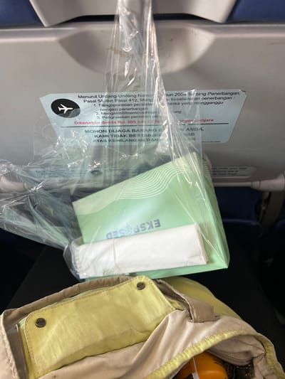 The image appears to be taken inside an airplane cabin. In the foreground, a yellow fabric bag with metal snap fasteners is visible. Attached to the seat in front of the bag is a clear plastic bag containing several items, including what looks like a green packet with white lines and a white folded item. A sticker is adhered to the seat, with text in Indonesian concerning regulations for passengers and penalties for violations. The background shows the back of an airplane seat and a blue headrest. The overall lighting suggests an indoor environment, likely during a flight. No specific location within Baji Mangngai, Indonesia can be determined from the visual information.