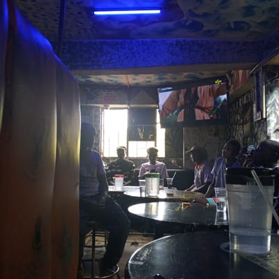 The image shows a group of men sitting around tables in what appears to be a bar or pub in Kinoo ward, Kenya. The location is indoors, with dim lighting enhanced by a blue light fixture on the ceiling. A television is mounted on the wall, displaying an image of a person. The men are seated around dark, round tables with clear pitchers and drinking glasses on them. One man is smoking. The environment is dimly lit with some daylight coming through windows in the background. The decor is textured, and there are warning signs posted on the wall. The image shows an ordinary social gathering in a relaxed setting.