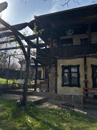 A building in Gorno Trape, Bulgaria, constructed from wood and plaster, features a prominent wooden framework. Two windows with dark frames are visible, with one on the first floor. A wooden balcony with a railing overhangs the ground level. A wooden pergola structure with climbing plants is in the foreground, and green grass is visible to the right.