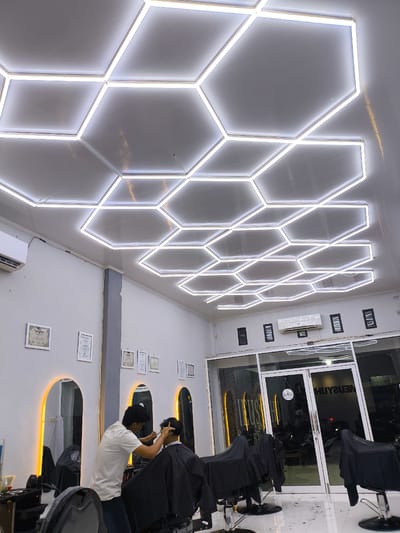 The image captures the brightly illuminated interior of a modern barbershop or hair salon located in Miruektaman, Indonesia. The ceiling is the prominent feature, covered entirely by an extensive network of interconnected hexagonal LED light panels emitting a cool white light. The walls are painted a light gray or white, adorned with multiple framed certificates or awards. On the left wall, an air conditioning unit is mounted near the ceiling. Further along the back wall, three arched mirrors are visible, each featuring a warm yellow backlighting strip, indicating styling stations.
In the midground, a male barber, wearing a light-colored t-shirt and dark trousers, is actively engaged in cutting the hair of a male customer. The customer is seated in a black barbershop chair, covered by a black cape, facing away from the camera. Several other black barbershop chairs, also draped with black capes, are arranged throughout the salon, some appearing unoccupied. Another air conditioning unit and three small, dark-framed rectangular wall fixtures are visible on the upper right side of the back wall. The front of the establishment is a large glass storefront with white-framed double doors. Reflections on the glass display reversed text, including "CLOSE" on a circular hanging sign, and blurred elements from the dark exterior, suggesting nighttime. The floor is tiled with light-colored (likely white) tiles, showing scattered hair clippings.