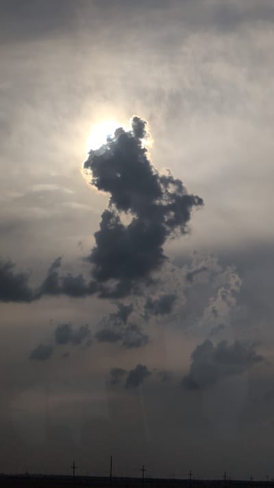 The image depicts a dramatic sky scene with the sun partially obscured by a dark, cumulus cloud formation. The sun's rays create a bright halo effect, illuminating the edges of the cloud. The foreground is a dark, silhouetted landscape, likely a field or plain, with faint outlines of utility poles or distant structures. The overall mood is somber and somewhat ominous, suggesting either an approaching storm or the aftermath of one. The location in Borcea, Romania, is not visually evident from the image itself, as the scene focuses solely on the sky and a featureless ground.